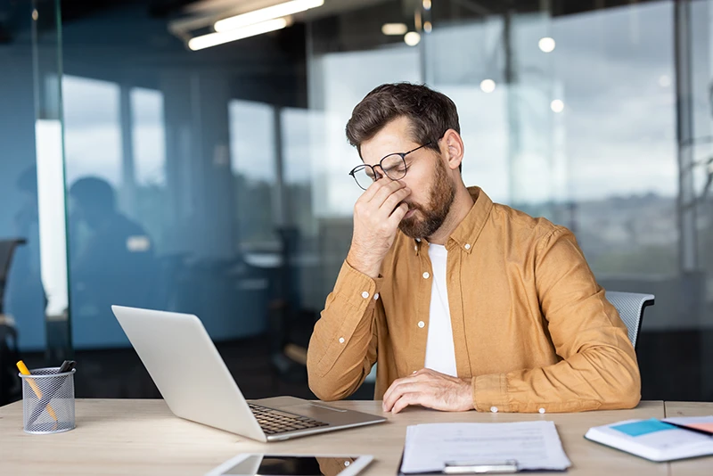 a man sitting at his desk at work and massaging the bridge of his nose due to a headache